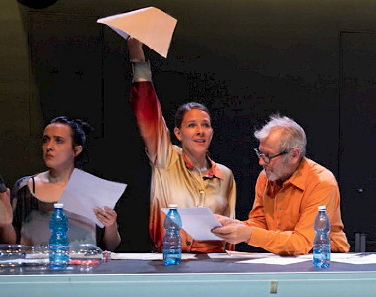 Mensch, du hast Recht! Im Bild stehen drei Personen an einem Sitzungstisch. Vor ihnen stehen 3 blaue PET-Flaschen und viele beschriebene A4-Blätter. Die Schauspielerin in der Mitte streckt mit der rechten Hand entschlossen eine weisse A4-Seite in die Höhe. Ihr Gesicht ist zuversichtlich. Sie trägt eine orange-beige Bluse und hat ihre mittelblonden langen Haare hinten zusammengebunden. Zu ihrer Linken beugt sich ein grauhaariger Mann mit orangem Hemd und Lesebrille über ein Dokument, das er in der linken Hand hält. Am linken Bildrand steht eine weitere Schauspielerin; sie trägt ein grauschwarzes Top. Ihre Haare sind schwarz und zu einem Dutt zusammengebunden. Auch sie hält in der Hand ein Blatt Papier und schaut konzentriert und kritisch in die Weite.